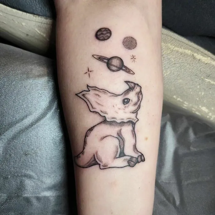 Tattoo of a dinosaur gazing at planets and stars, capturing outer space cool vibes.