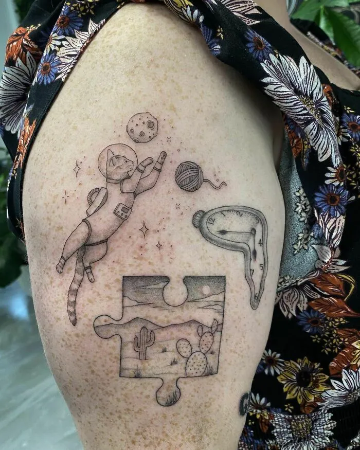 Cat astronaut tattoo with space-themed elements including a planet, clock, and landscape puzzle piece on an arm.