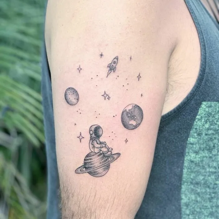 Astronaut sitting on Saturn tattoo, featuring planets and a rocket, perfect for astrology enthusiasts.