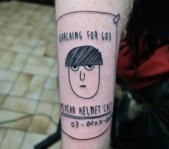 Tattoo of cartoon face with text "Searching for God" and "Psycho Helmet Cult," embodying unique word tattoo design.