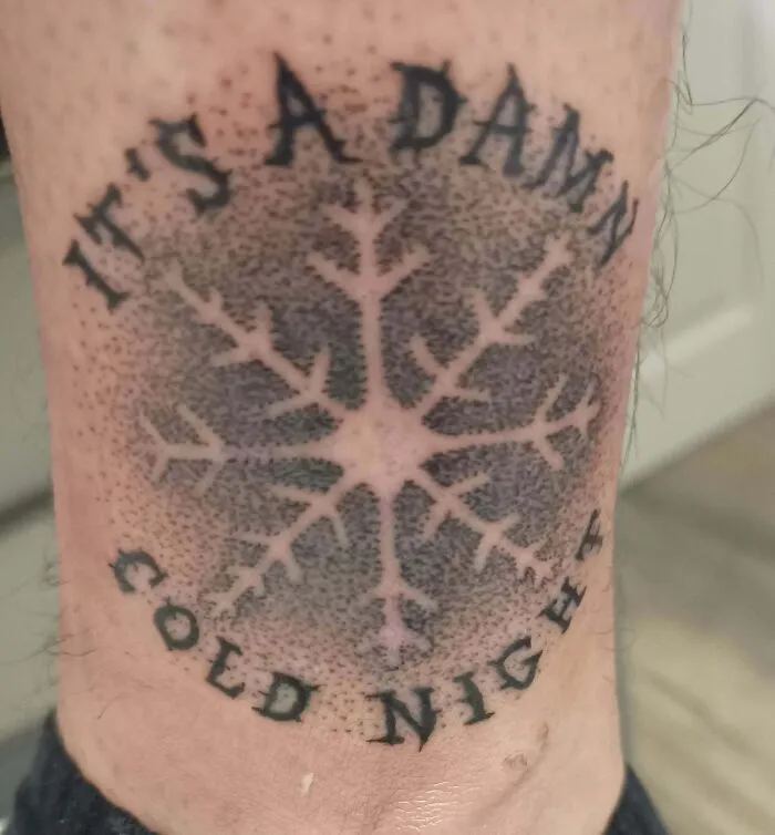 Tattoo of a snowflake with the words "It's a damn cold night," featuring intricate dotwork and bold lettering.
