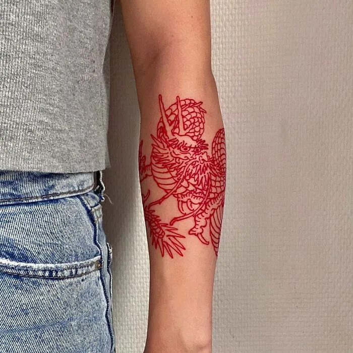 Red ink tattoo of a detailed dragon design on forearm wearing a gray shirt and denim jeans.