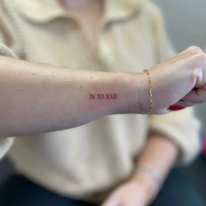 Red ink tattoo on forearm with Roman numerals IV XII XXII, close-up shot.