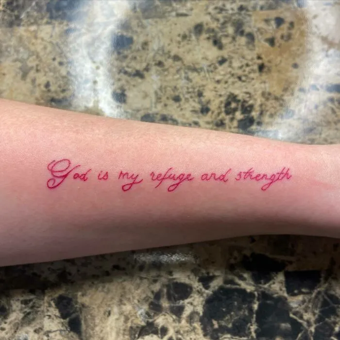 Red ink tattoo on forearm reading "God is my refuge and strength" in elegant script.