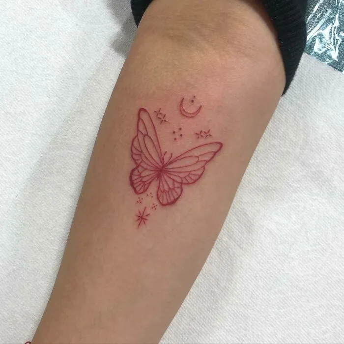 Red ink butterfly tattoo with crescent moon and stars on forearm.