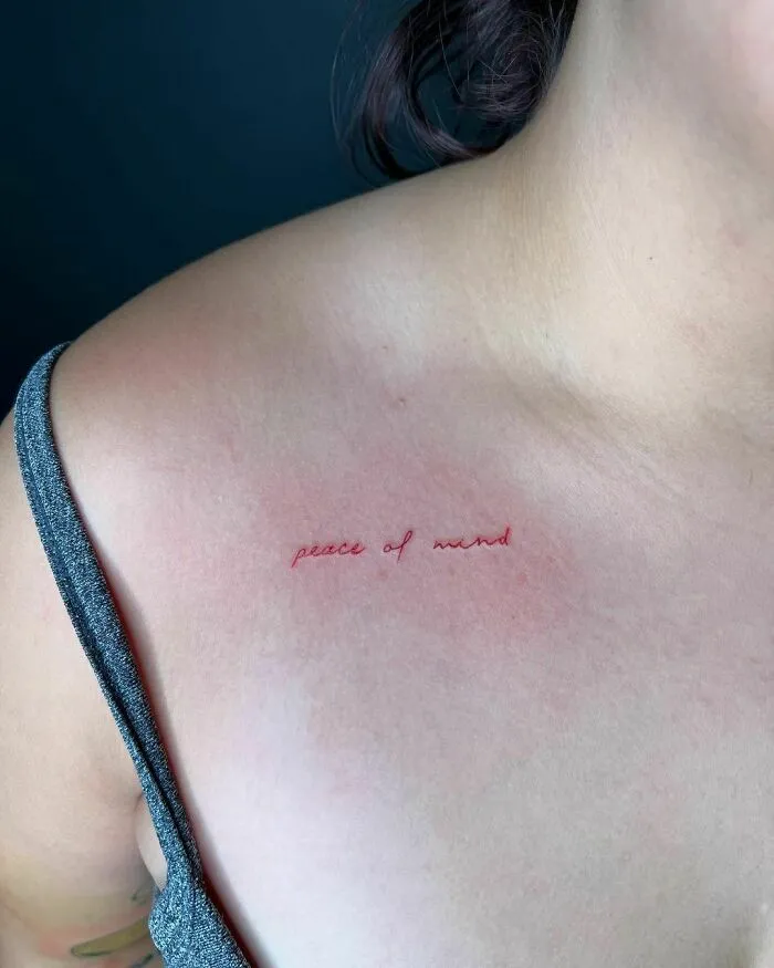 Red ink tattoo on collarbone with "peace of mind" text, showcasing minimalist design.