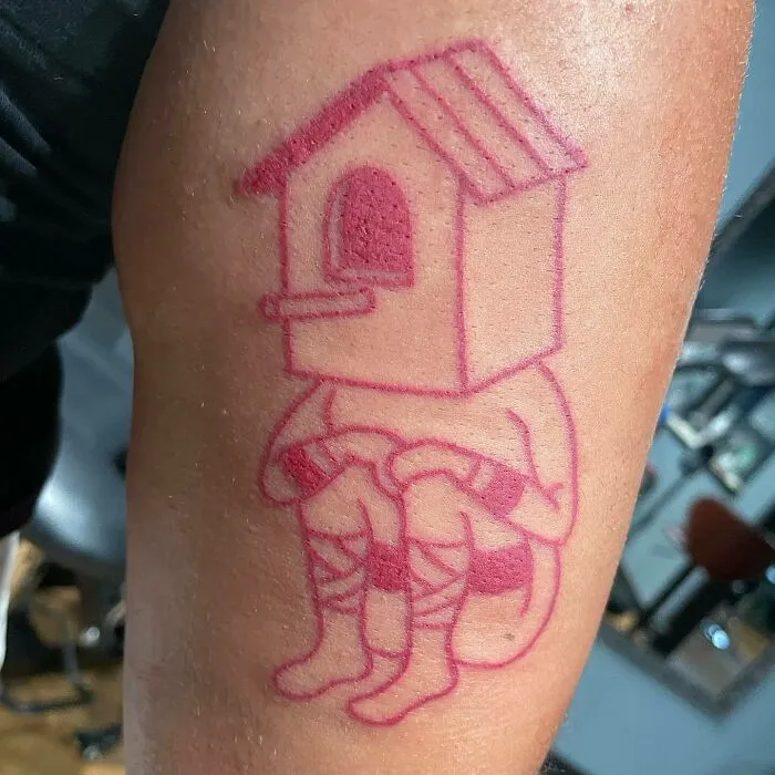 Red ink tattoo of a person with a birdhouse head, seated with knees drawn up, showcasing unique design on skin.
