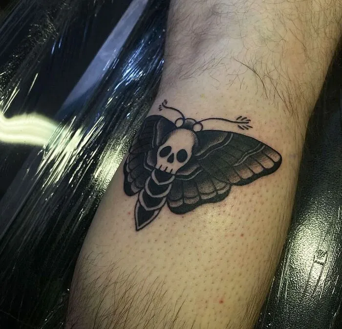 Moth tattoo with a skull design on the lower leg, showcasing bold lines and dark shading.