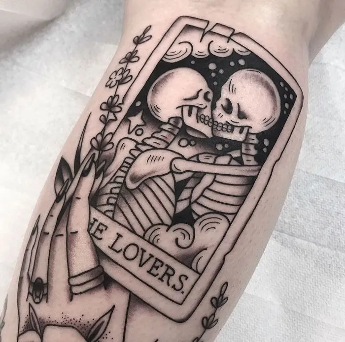 Lower leg tattoo featuring skeletons in an embrace within a tarot card labeled "The Lovers."