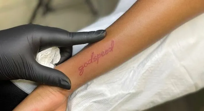 Red ink tattoo on forearm reading "godspeed," with artist wearing black gloves.