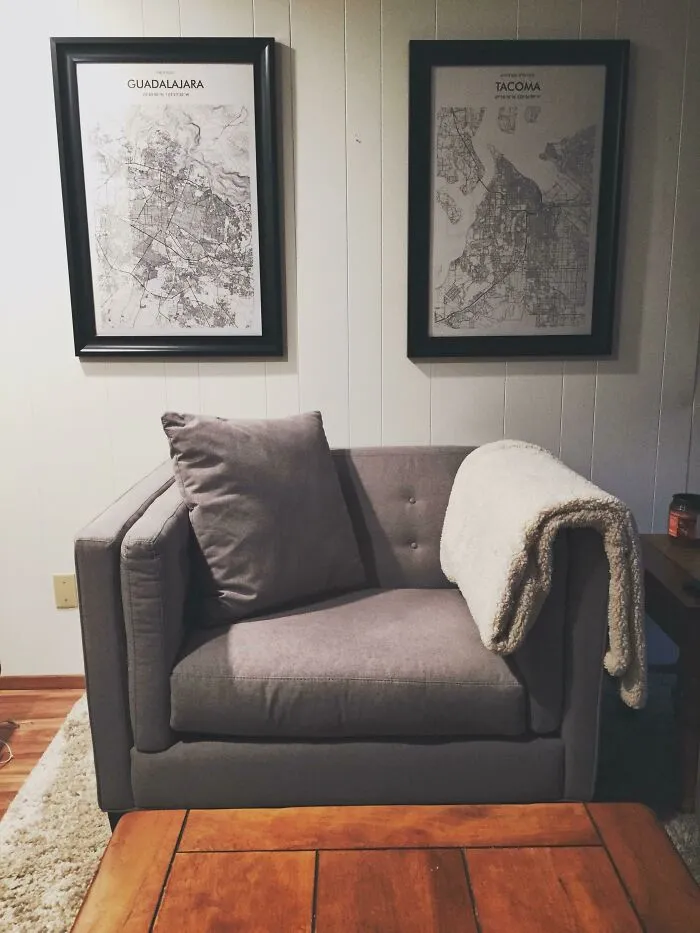 Gray armchair with a cozy blanket, flanked by framed city maps on the wall, enhancing home decoration elegance.