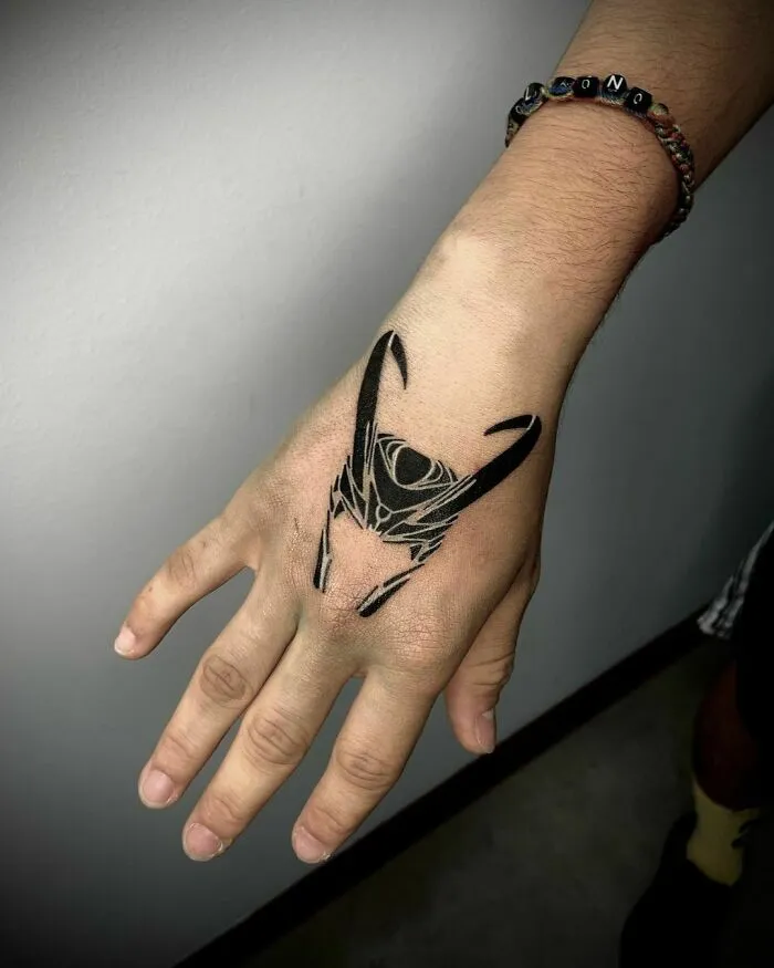 Hand featuring a detailed Marvel tattoo of Loki's helmet, emphasizing line work and bold black ink.