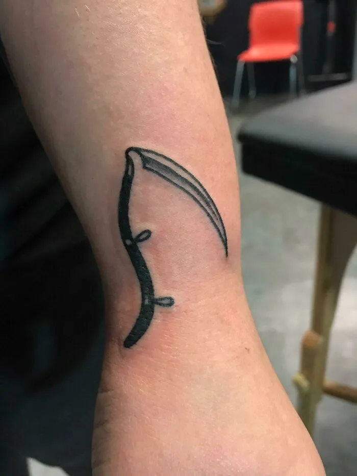 Minimalist scythe wrist tattoo design on forearm with black ink.