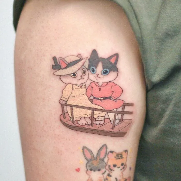 Tattoo of two cute cartoon cats on a person's arm, animal-themed design by a tattoo artist.
