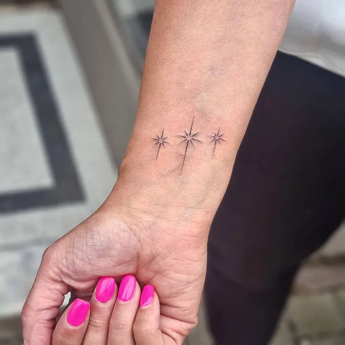 Wrist tattoo featuring three small star designs with a geometric style, displayed on a hand with pink-painted nails.