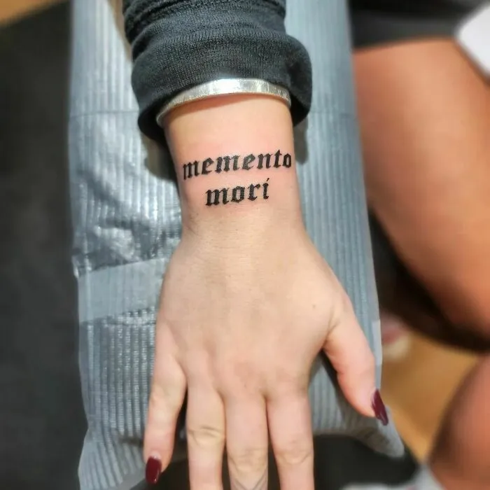 Wrist tattoo design with "memento mori" text in bold black script.