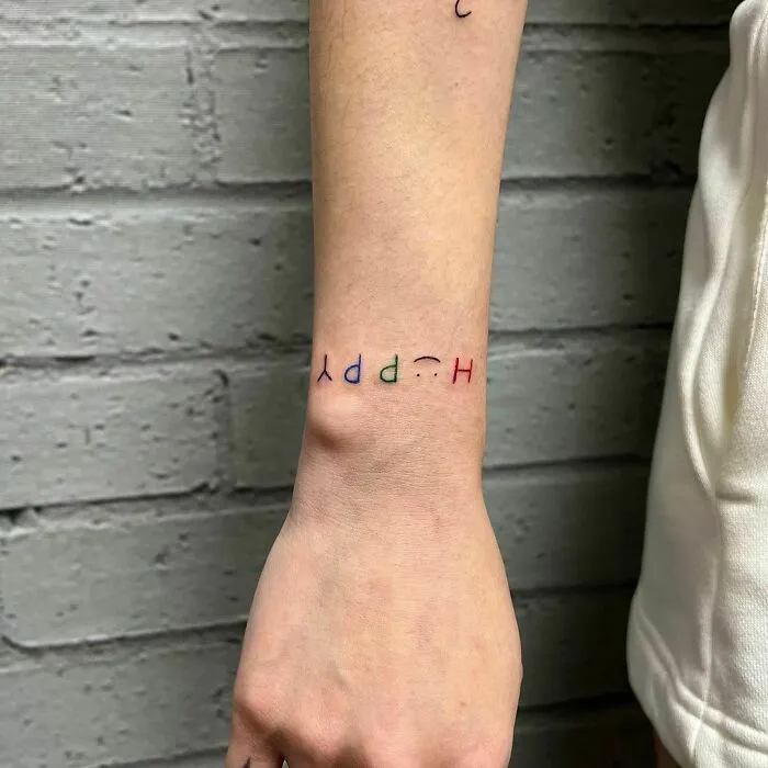 Colorful wrist tattoo design reading "happy" on a person's arm against a brick wall background.