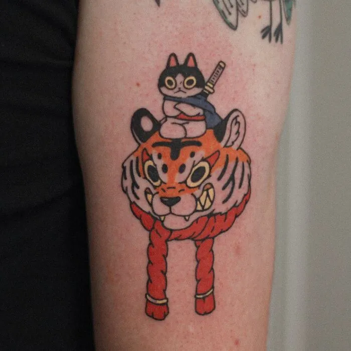 Tattoo of a cute cat with a sword sitting atop a tiger's head, showcasing animal-themed design.