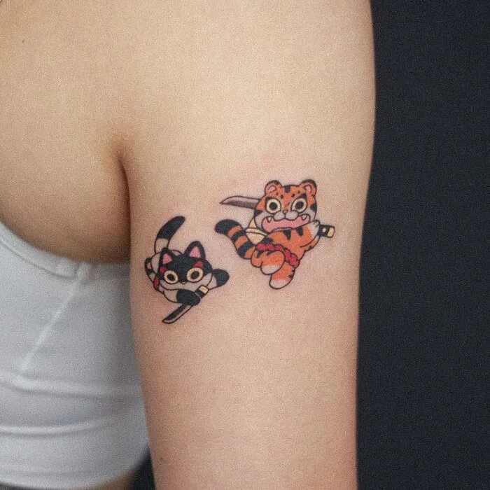 Tattoo of adorable animal-themed designs featuring a cartoon cat and tiger on an upper arm.