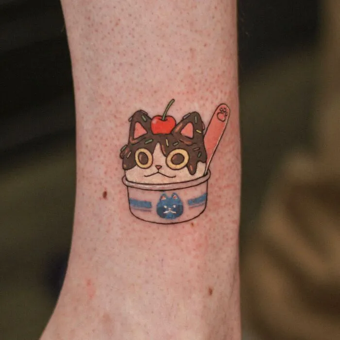 Adorable animal-themed tattoo of a cat in an ice cream bowl with a cherry on top, featuring vibrant colors.