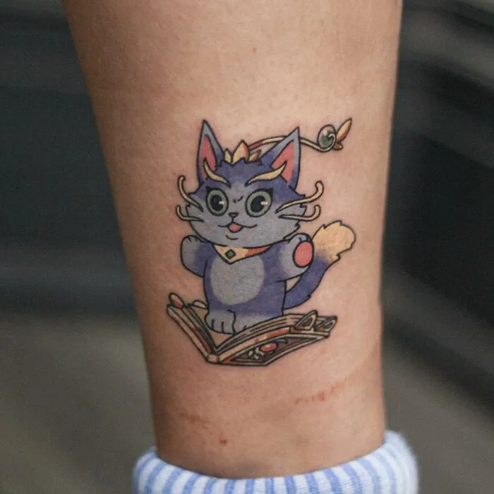 Adorable animal-themed tattoo of a whimsical cartoon cat holding a book on a person's forearm.