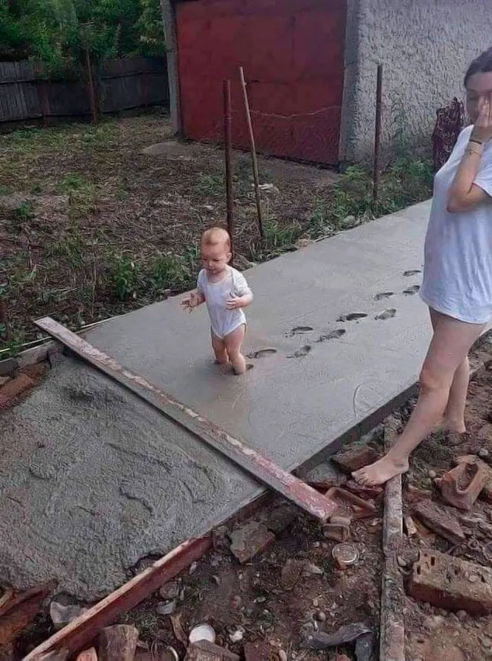 Baby walking on wet cement, leaving footprints, with a woman nearby. Real unhinged image capturing a surprising moment.