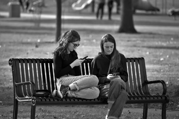 Two millennials on a bench using smartphones, embodying millennial slang culture.