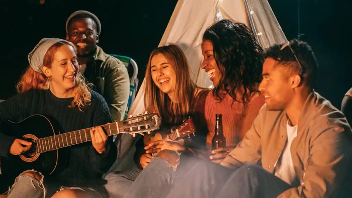 Millennials enjoying a campfire, playing guitar and laughing, in a casual outdoor setting.