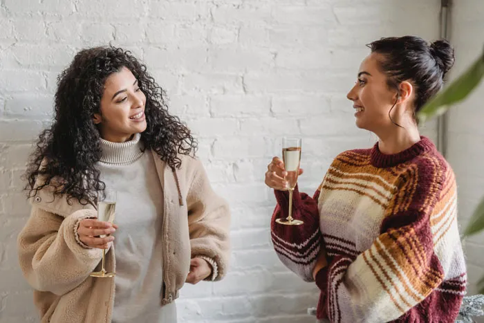 Two women in cozy sweaters converse, holding drinks, embodying millennial slang culture against a white brick wall.