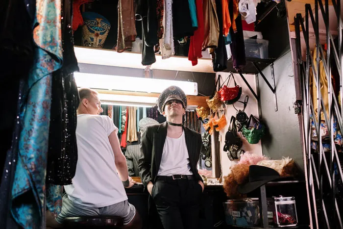 Two people in a dressing room trying on outfits, surrounded by colorful costumes, embodying millennial slang culture.