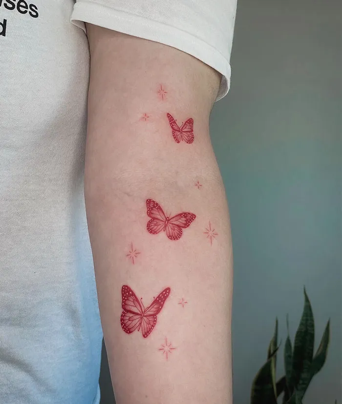 Red ink butterfly tattoos on an arm, featuring delicate, artistic detailing with small star accents.
