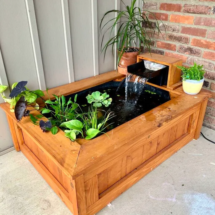 Wooden aquarium with plants and a small waterfall, set against a brick wall.
