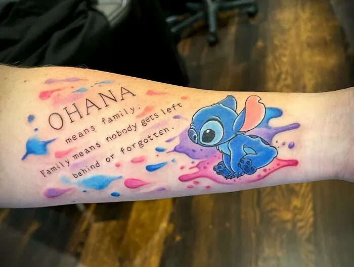 Tattoo of "Ohana means family" with colorful splashes and a cartoon character, symbolizing unbreakable family bonds.
