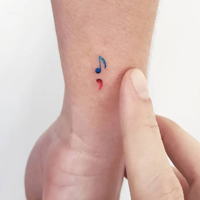 Small music tattoo with a blue note and red semicolon on wrist.