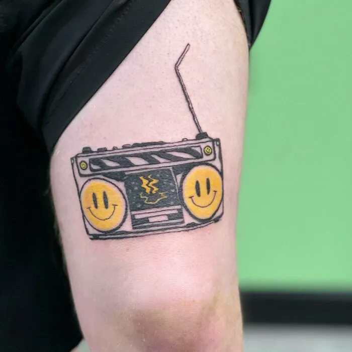 Tattoo of a retro boombox with yellow smiley faces, symbolizing music on an upper arm with a green background.