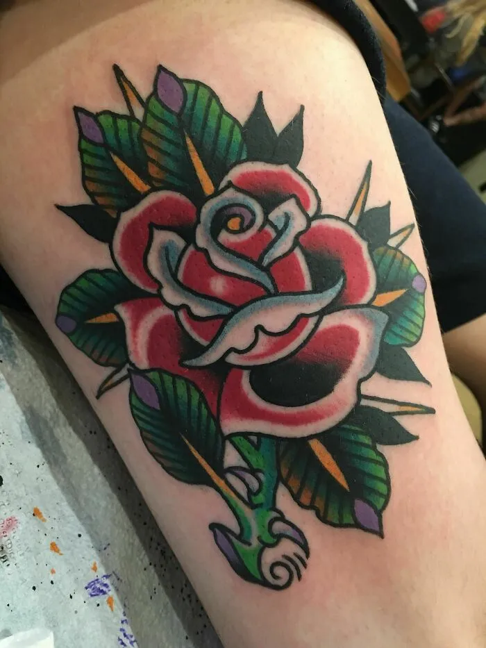 American traditional tattoo of a vibrant red rose with green leaves on skin.