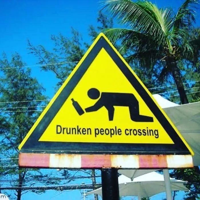 Funny sign showing a stick figure crawling with a bottle, labeled "Drunken people crossing," against a tropical background.