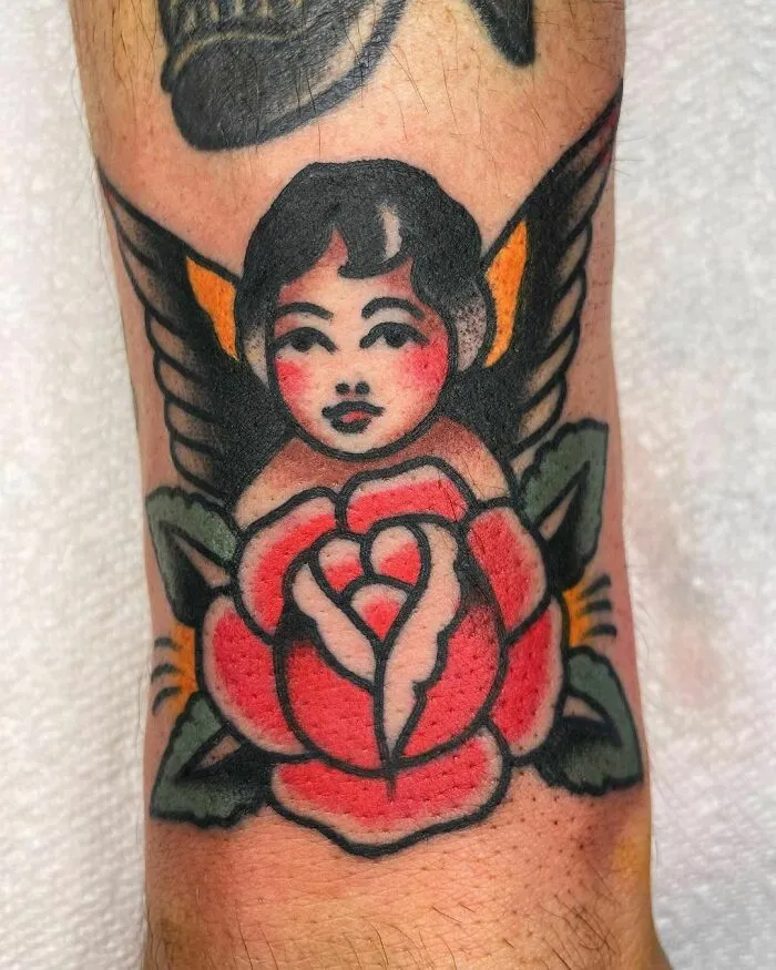 American traditional tattoo of a winged cherub above a red rose on skin.