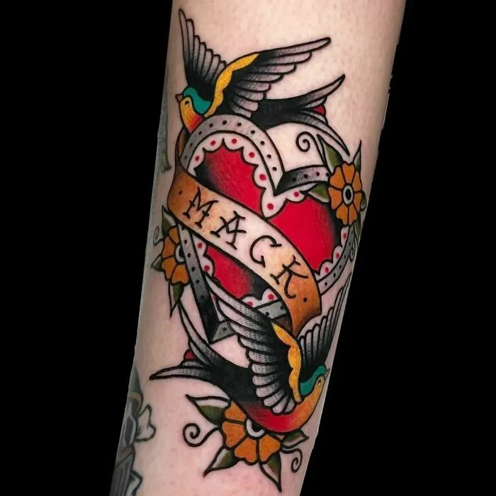 American traditional tattoo with a heart, banner, swallows, and flowers on an arm.