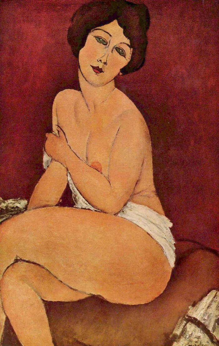 Nude Sitting On A Divan by Amedeo Modigliani, 1917, oil painting depicting a partially draped woman seated with crossed legs against a warm red background
