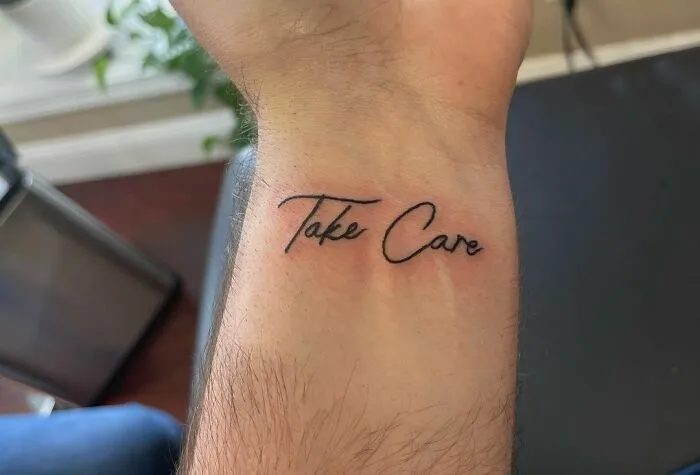 Minimalist wrist tattoo design with the words "Take Care" in elegant script.