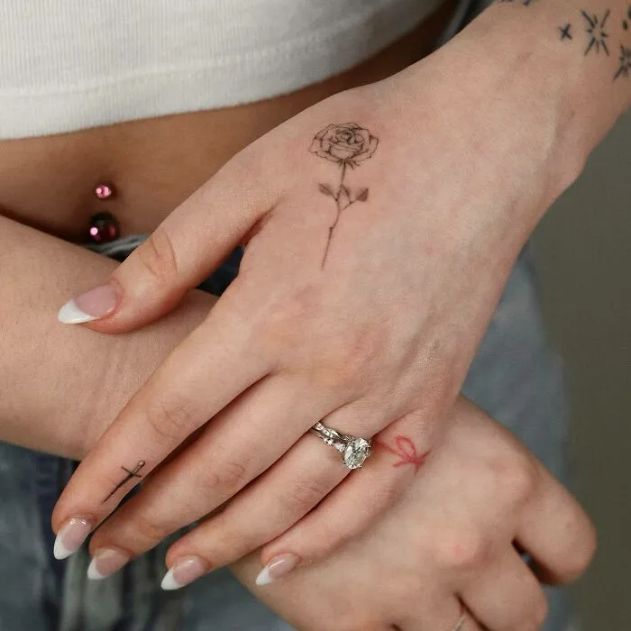 Close-up of small hand tattoos featuring a delicate rose, cross, and ribbon design.
