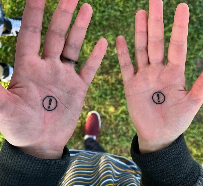 Small hand tattoos featuring exclamation marks inside circles on palms, viewed outdoors with striped clothing.