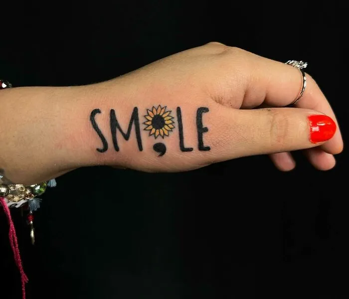 Small hand tattoo with the word "SMILE" using a sunflower for the "O" and vibrant red nail polish.