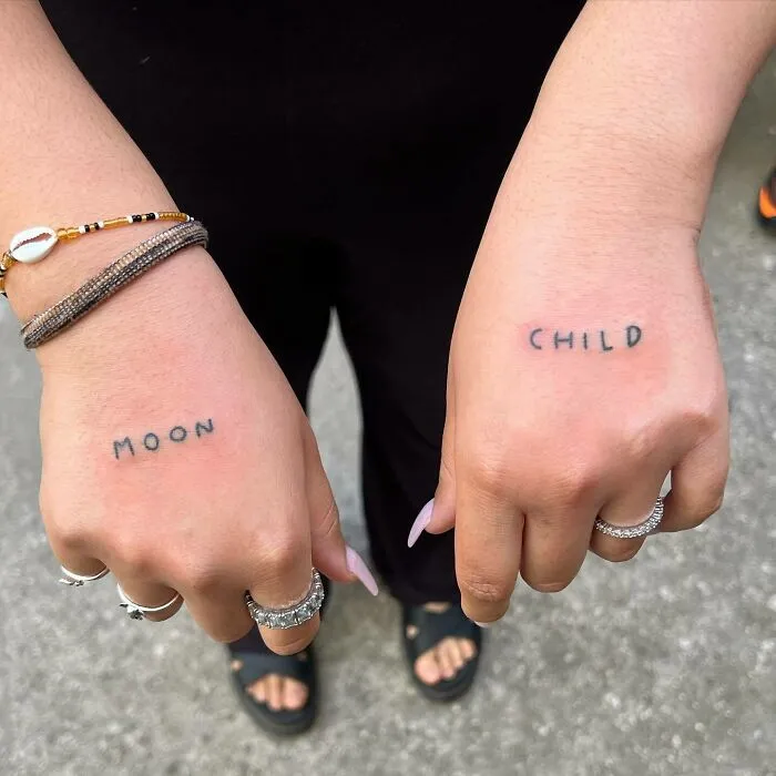Small hand tattoos reading "moon" and "child" on the knuckles, with jewelry on fingers, showcasing minimalist design.