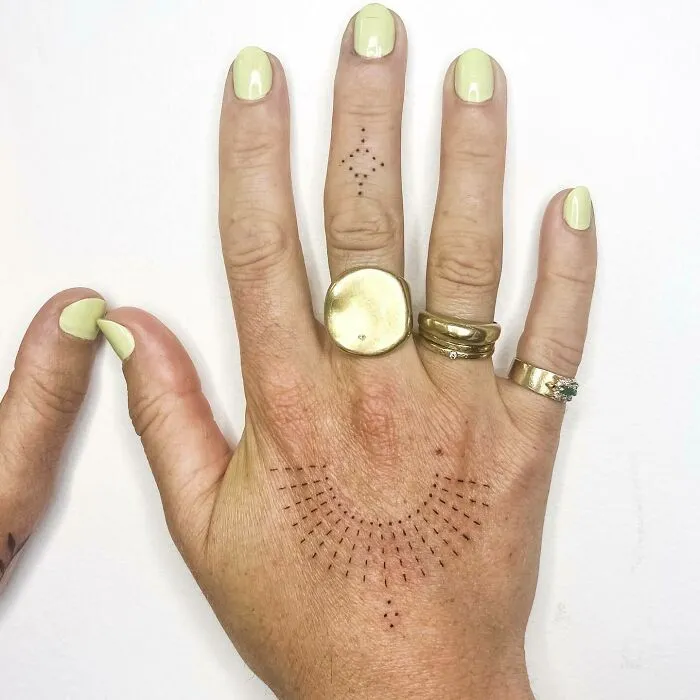 Hand showcasing elegant small tattoos with geometric patterns, complemented by gold rings on manicured fingers.