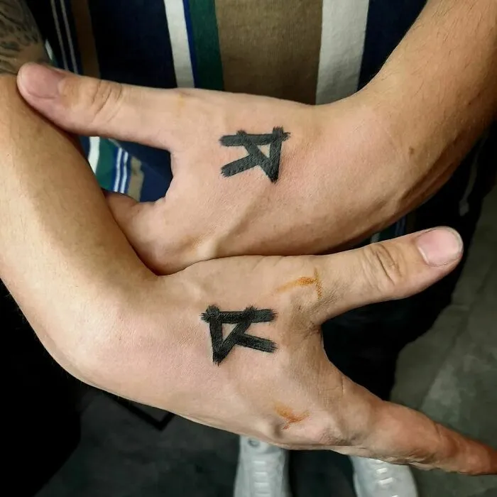 Two small hand tattoos featuring black symbols on the thumbs and webbing of intertwined hands.