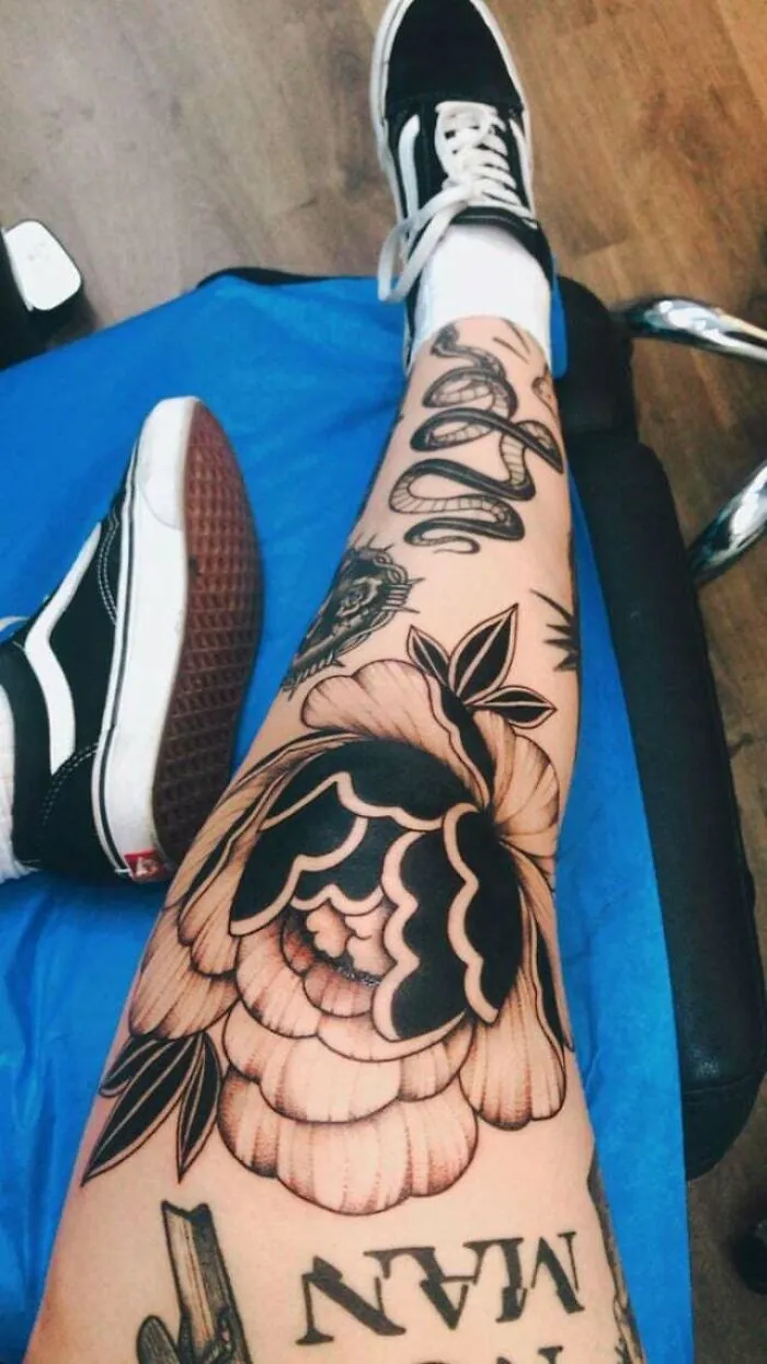 Leg showcasing knee tattoos with floral and snake designs, wearing Vans shoes, on a blue cushion in a tattoo studio.