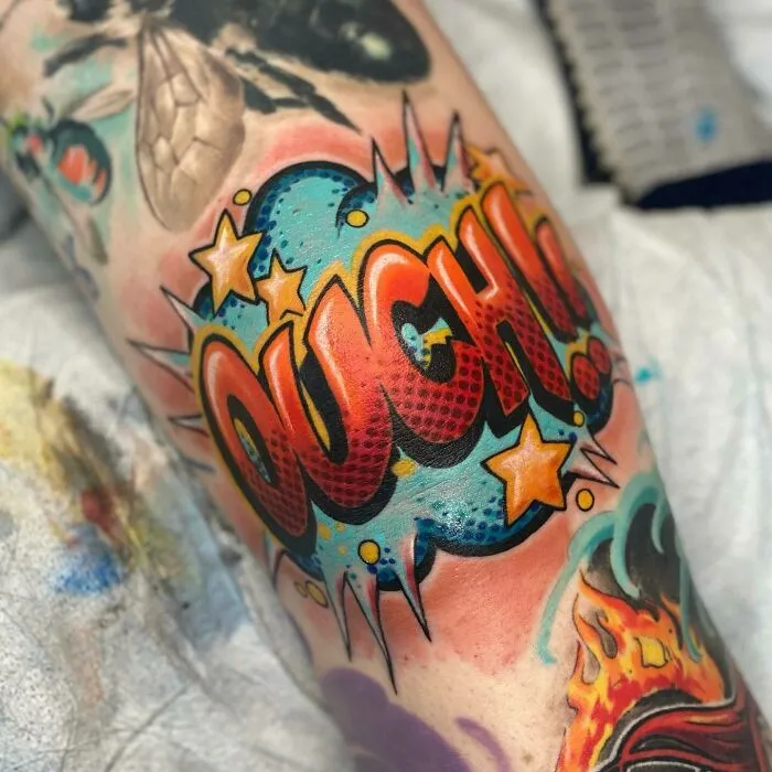 Colorful comic-style knee tattoo with the word "OUCH" in bold letters and vibrant stars.