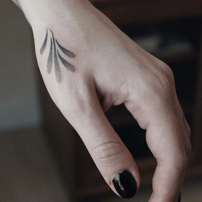 Black and gray small hand tattoo with abstract design on the back of the hand, black nail polish visible.